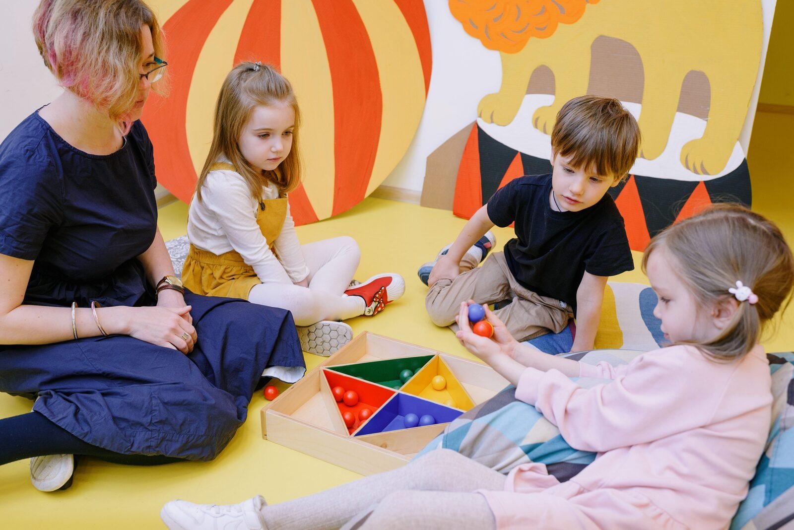 An educator and preschool children playing with colourful learning materials in a bright childcare classroom.