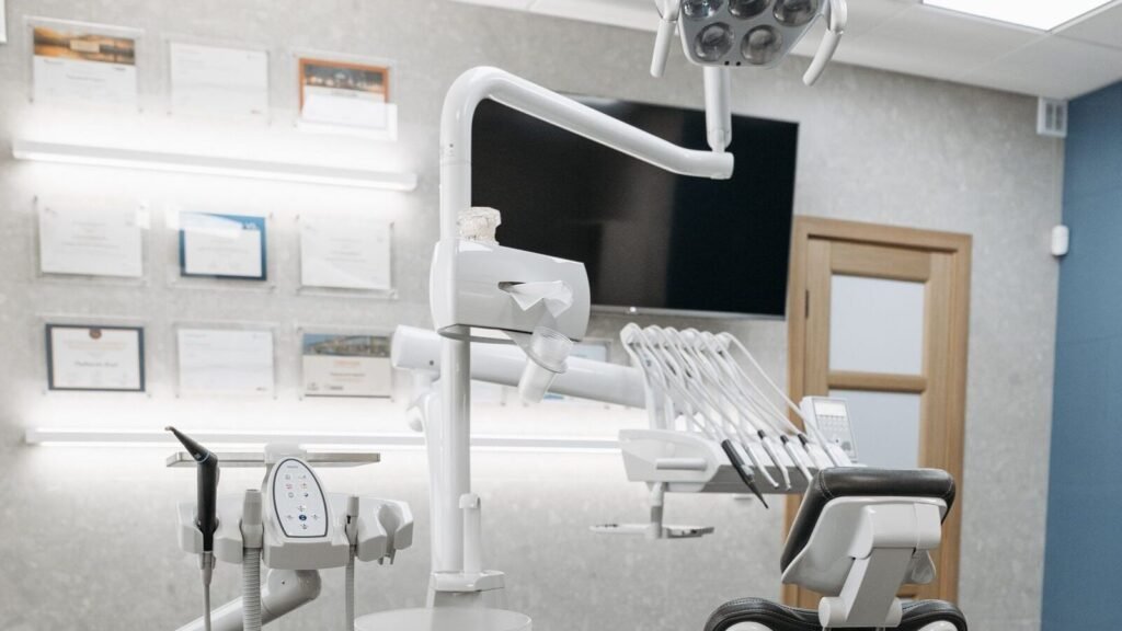 Empty modern dental treatment room with dental chair, overhead light and surrounding clinical surfaces.